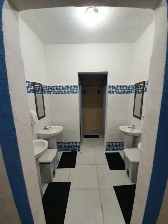 Banheiro com paredes de azulejo branco e detalhes em azul, duas pias no lado esquerdo, duas pias no lado direito, pequeno espelho, tapete e lixo em ambos os lados também.