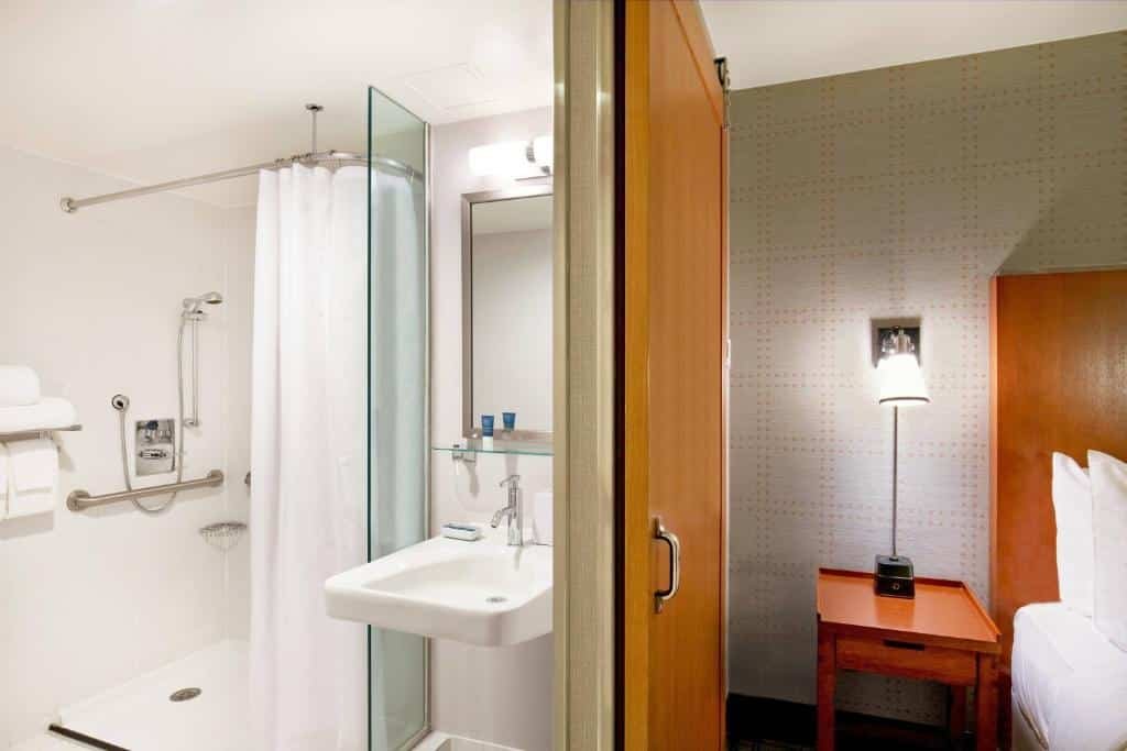 dois ambientes divididos por uma porta, mostrando do lado direito uma parte da cama de casal da suíte e do lado esquerdo o banheiro adaptado para pessoas com deficiência motora. Ou seja, há barras de apoio no box e uma pia vazada e rebaixa para atender a demanda de cadeirantes.