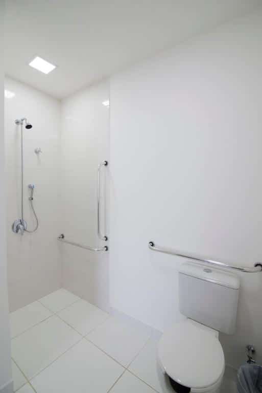 banheiro do Hotel H2 Congonhas com barras de apoio dentro do boxe de banho e ao redor do vaso sanitário. O espaço é todo branco, tanto nos azulejos e parede, quanto nas porcelanas