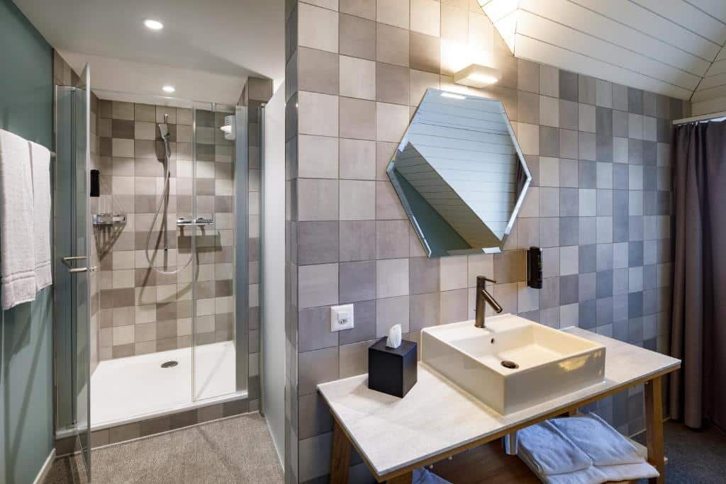 Banheiro moderno no hotel Münzgasse. Na direita vemos um gabinete de pia com porcelanato e um espelho hexagonal. Atrás, vemos o box do banheiro com a ducha.