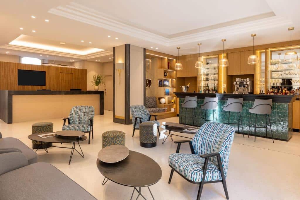 Lounge do Hôtel Apollinaire Nice. Nos fundos há o balcão de atendimento do hotel, no lado direito há um bar elegante com quatro banquetas em frente ao balcão do bar e atrás há diversas cadeiras e mesas de centro.