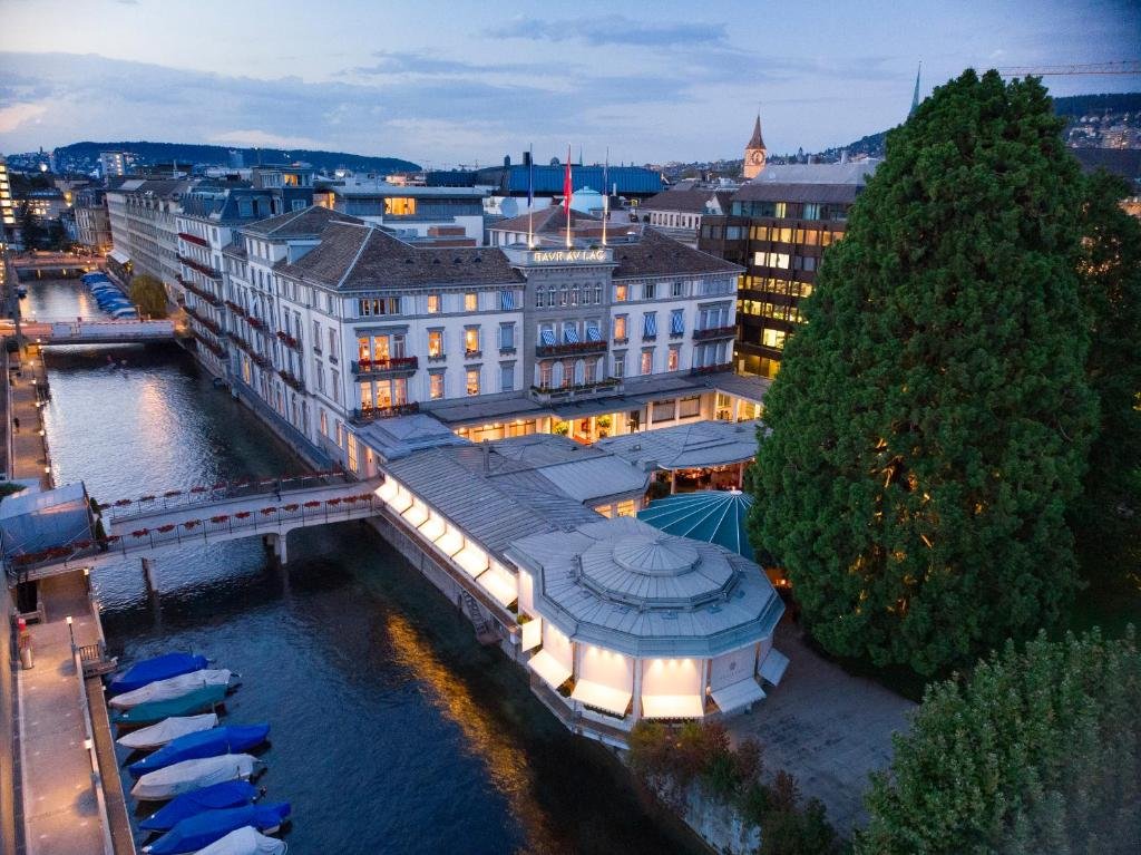 Prédio do Baur au Lac, um dos hotéis em Zurique, no início da noite com o céu começando a escurecer, com luzes acesas, ao lado de um rio, e uma árvore grande verde na frente