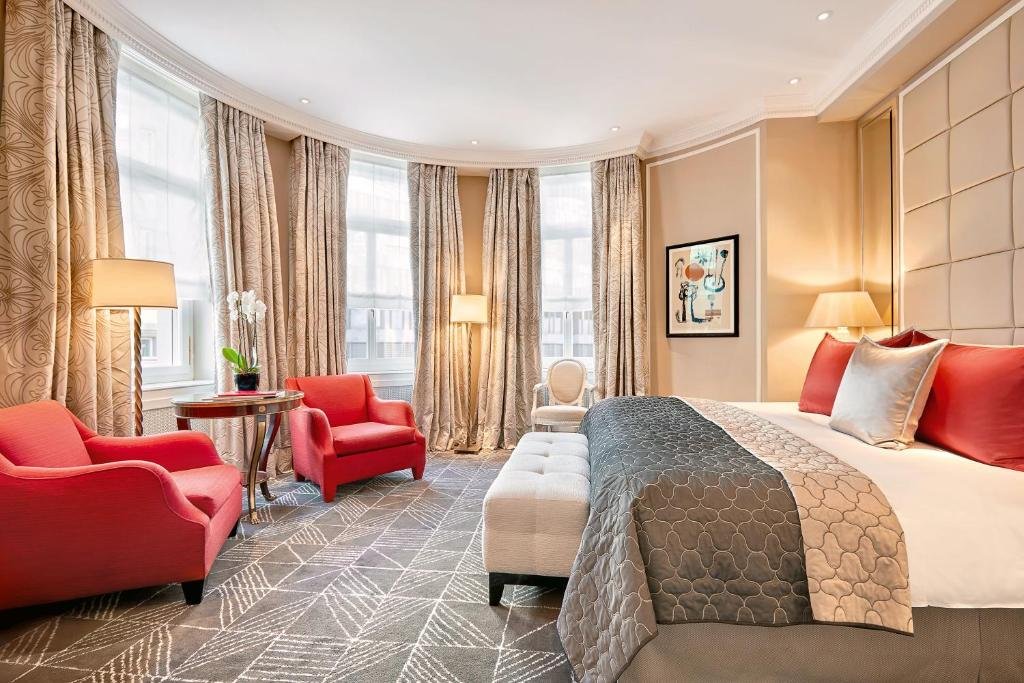 Quarto do Baur au Lac, com decoração elegante composta por duas poltronas vermelhas, janelas de vidro com várias cortinas altas, mesa redonda com flor e cama com almofadas em cima