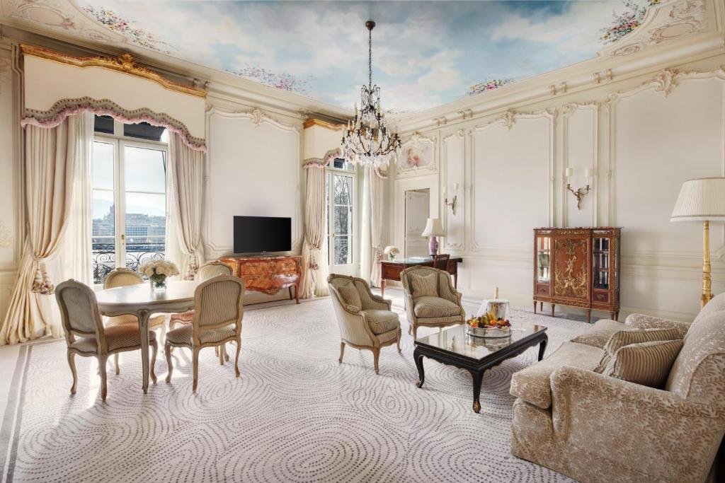 Outro ângulo de uma suíte no Beau Rivage Geneve. No canto inferior direito, há um sofá claro, uma mesa e centro na frente e duas poltronas da mesma cor. Do outro lado do sofá, encostado na parede no fundo, há um abajur e uma estante marrom. Já no lado esquerdo, há uma mesa redonda de jantar com quatro cadeiras. No fundo do quarto, no centro na parede, há uma mesa de TV, com televisão em cima. Há duas janelas no lado direito e esquerdo da TV. O teto é pintado como se fosse um céu claro com nuvens e possui um lustre.