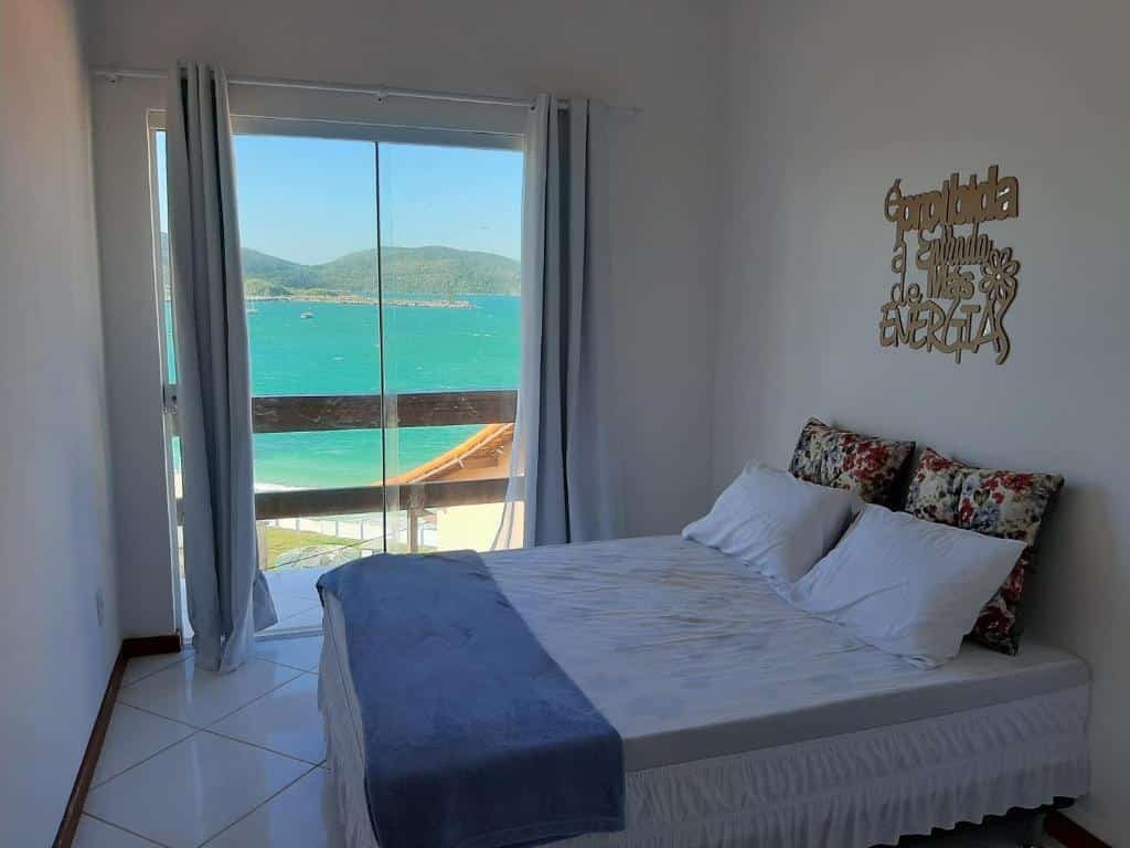 Quarto do Bela Vista Flats. No lado direito do quarto a cama de casal está encostada na parede e no fundo há uma porta de vidro com cortinas ao lado que dá acesso a varanda e oferece vista para o mar. Imagem utilizada para ilustrar o post airbnb em Arraial do Cabo.