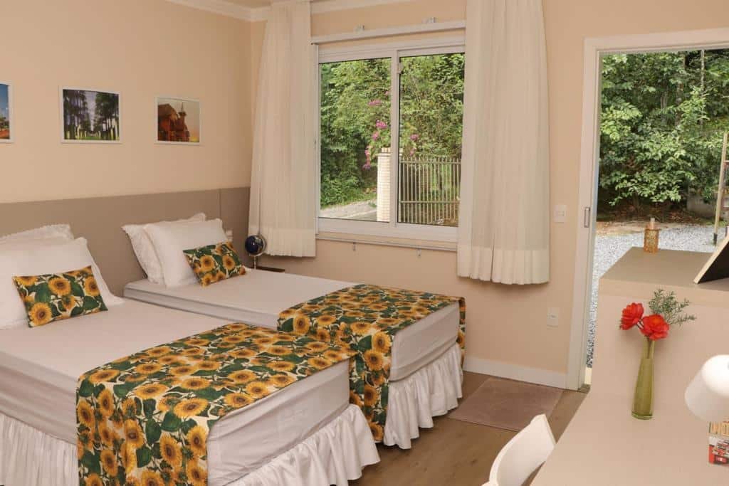 quarto da Bela Vista Hospedagem com duas camas de solteiro dispostas lado a lado no canto esquerdo da imagem. Em frente às camas está uma cômoda de madeira com mesa e cadeira para trabalho. Ao fundo do quarto é possível ver a janela e a porta, uma do lado da outra, ambas abertas e com vista para uma área com muitas árvores verdes.