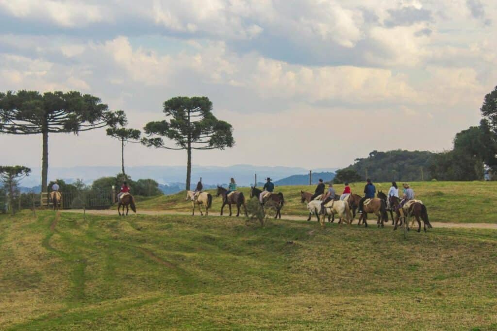 Um grupo de pessoas cavalgando por uma trilha em uma colina gramada no Boqueirão Hotel Fazenda, com árvores altas e um céu nublado ao fundo.