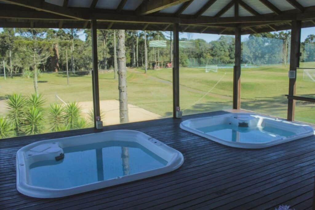 Duas banheiras de hidromassagem quadradas em um deck de madeira sob um gazebo, com vista para um campo de golfe com árvores ao fundo. Esse é o Boqueirão Hotel Fazenda, um dos hotéis fazenda em Santa Catarina.