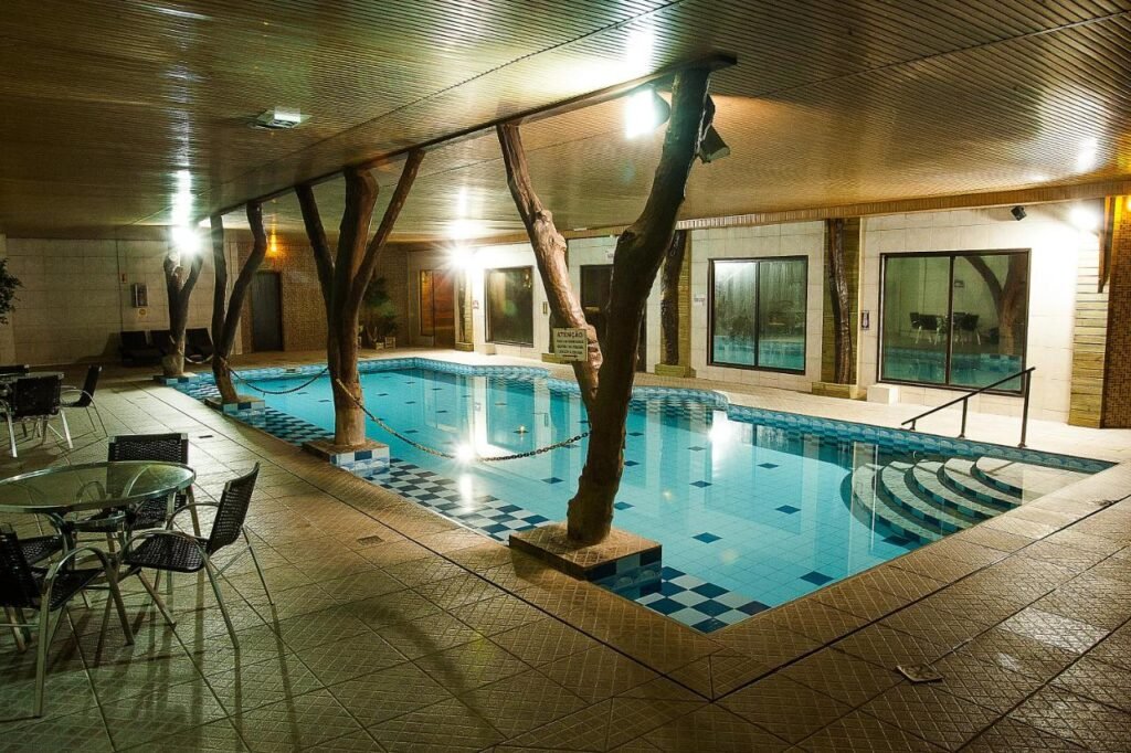 Área de piscina no Boqueirão Hotel Fazenda coberta com pilares de vigas de madeira, piscina com azulejos xadrez, mesas e cadeiras ao redor e grandes janelas em uma parede.
