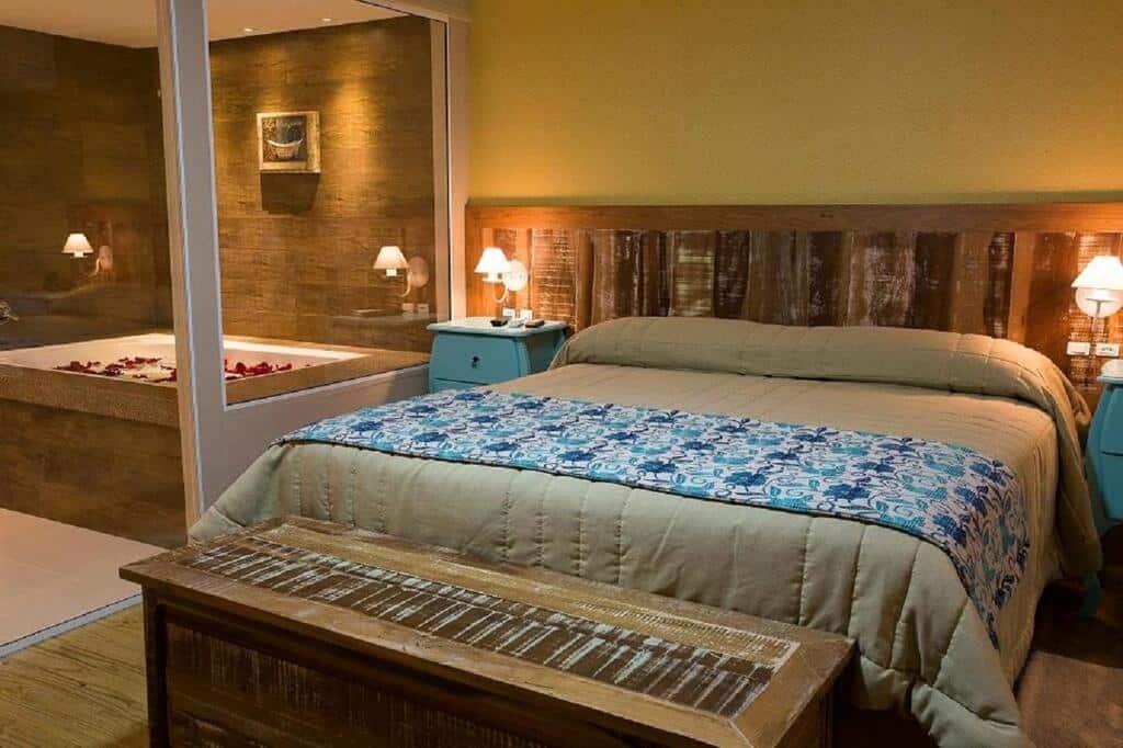 Quarto aconchegante com cama grande, cabeceira de madeira, mesa de cabeceira e banheiro visível com jacuzzi atrás de paredes de vidro. Esse é o Boqueirão Hotel Fazenda, um dos hotéis fazenda em Santa Catarina.