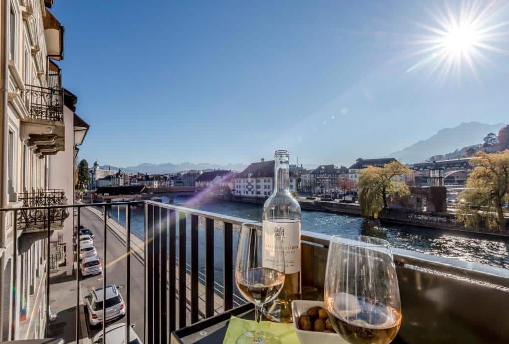 Vista da varanda do Boutique Hotel KARL. Vemos duas taças e uma garrafa de vidro com bebida. Há um rio passando em frente ao hotel. O sol brilha no céu. Carros estão estacionado na rua e podemos ver casas e construções de Lucerna.