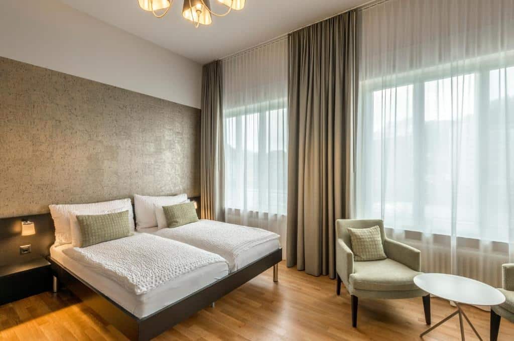 Foto do Boutique Hotel KARL, para representar o post de hotéis em Lucerna. O quarto é espaçoso e há uma cama box de casal na esquerda. No canto direito, há uma mesa redonda com duas poltronas em volta. Do lado direito da cama há duas grandes janelas que iluminam a suíte.