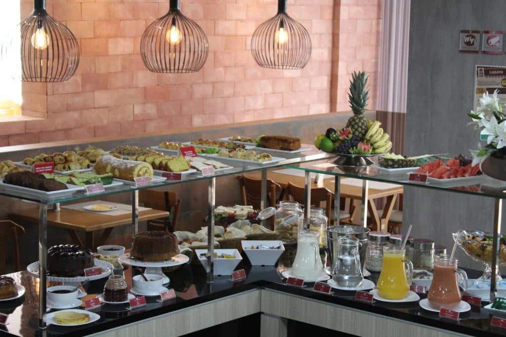 Área de refeições do Bristol Viçosa Hotel. Na frente da imagem há uma mesa em formato de L com diversas comidas e bebidas. Já no fundo estão as mesas com cadeiras.