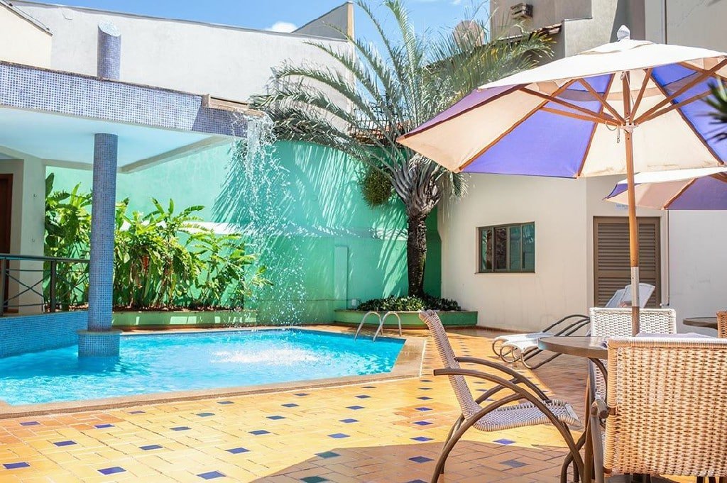 Piscina com mesa, cadeiras e guarda-sol ao lado. É possível ver árvore, algumas plantas e acomodações no canto. Foto para ilustrar post sobre hotéis em Campo Grande.