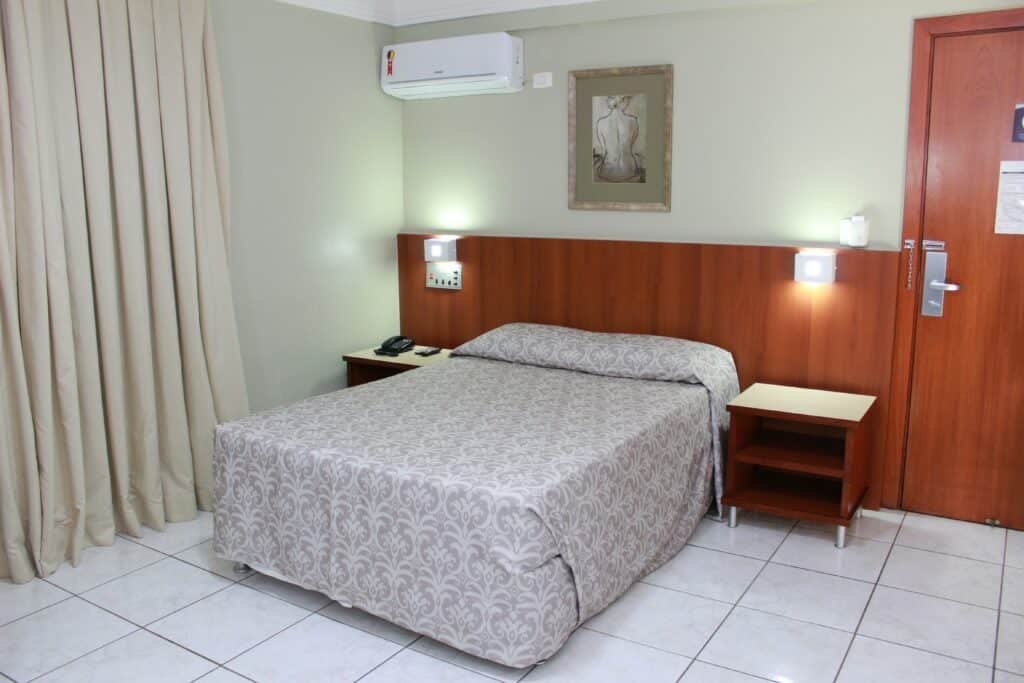 Quarto com uma cama de casal, luminárias e duas mesinhas aos lados. No canto é possível ver uma cortina.