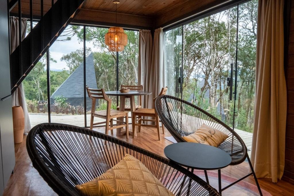 Interior das Cabanas Cold Mountain com mesas e cadeiras modernas, de estilos variados, baixos e altos, com janelas grandes de vidro por todos os lados, com vista para natureza.