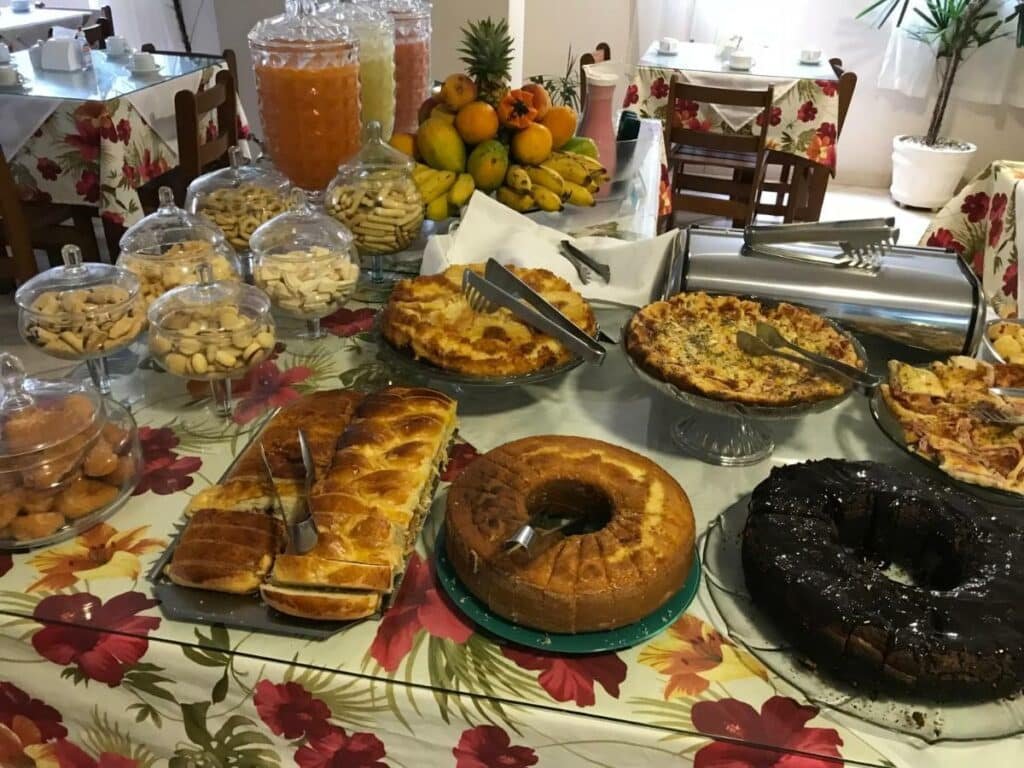 Mesa do café da manhã do Hotel Marlin Azul. Uma variedade grande de bolos, pães, sucos, frutas e bolachas. Ao redor, mesas com xícaras de café em cima.