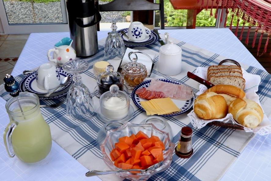 Café da manhã da Hospedagem Bela Vista, uma das opções das pousadas em Joinville, com pães, frios e frutas picadas em cima de belos pratos detalhados em azul. Há também xícaras e jarras de suco, além de uma garrafa térmica com café.