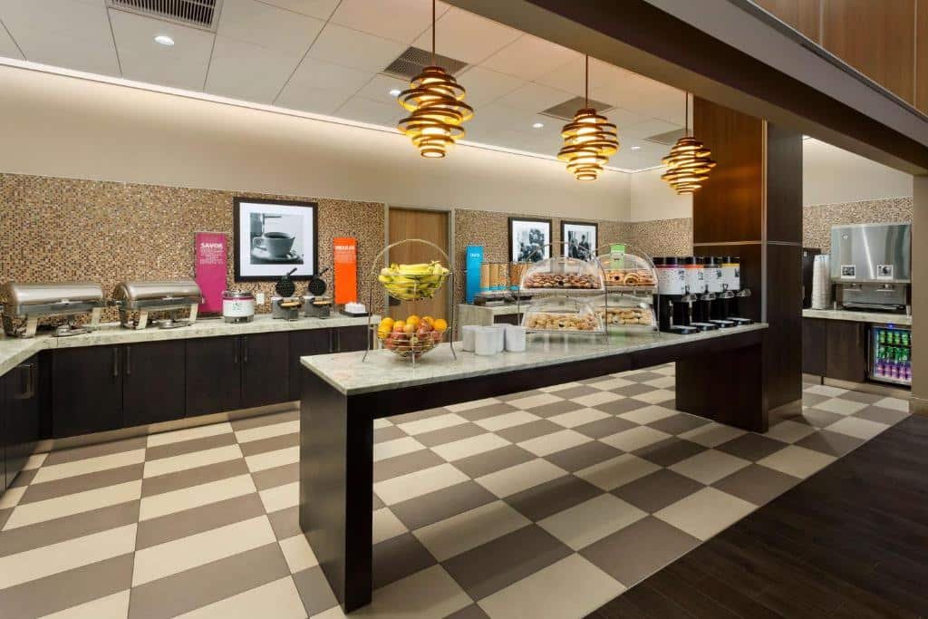 refeitório do Hampton Inn Times Square Central com um balcão central repleto de frutas e pães e bagels. Ao fundo há outros balcões de canto com waffles e pratos quentes.