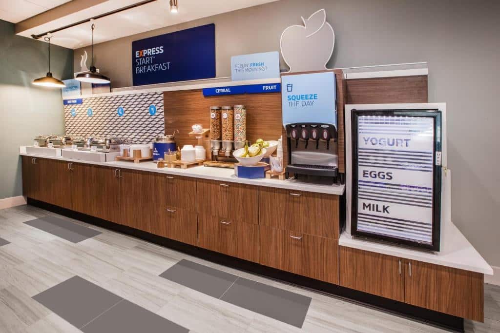 Imagem de um café da manhã no Holiday Inn Express Newark Airport. O buffet apresenta uma variedade de opções gastronômicas, incluindo frutas frescas, cereais, pães, sucos e café. As mesas estão dispostas de maneira organizada, com cadeiras ao redor, prontas para os hóspedes se sentarem e desfrutarem da refeição. O ambiente é iluminado e convidativo. O café da manhã oferece uma seleção ampla de alimentos para iniciar o dia de forma nutritiva.