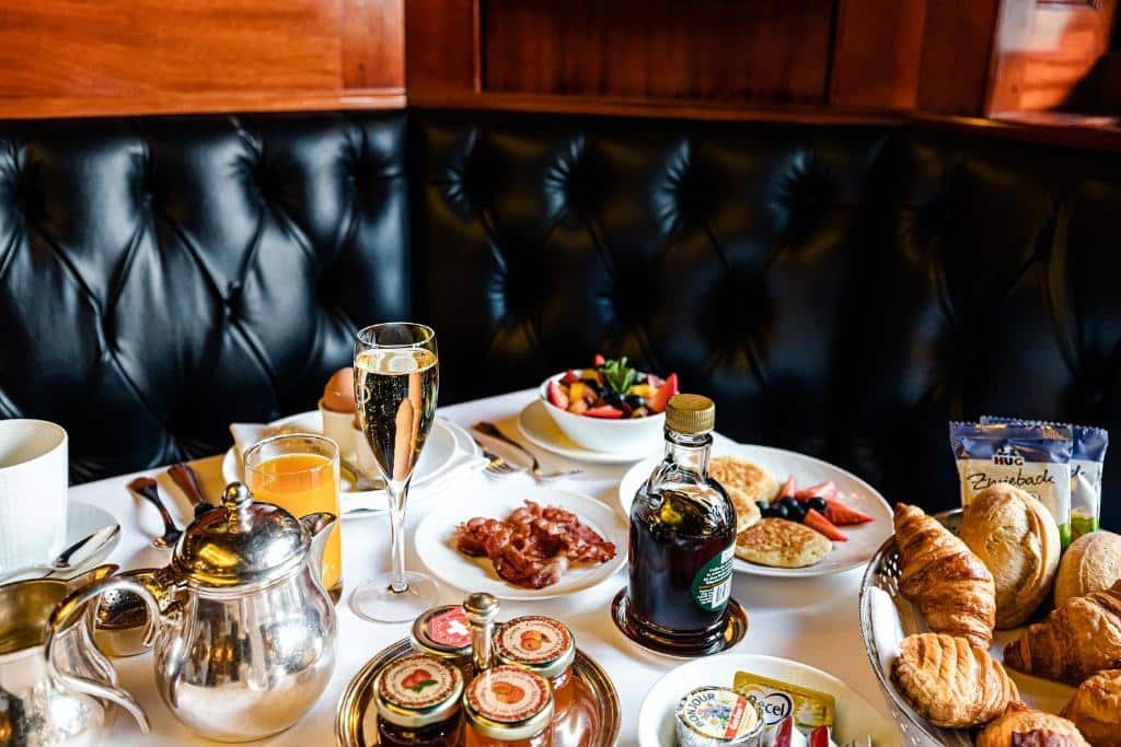 Um buffet de café da manhã no Hotel de la Cignone. A mesa é branca e está recheada de opções como geleias, pães, panquecas, frutas, taças, copo com suco, bule, etc. Atrás, há um sofá preto.