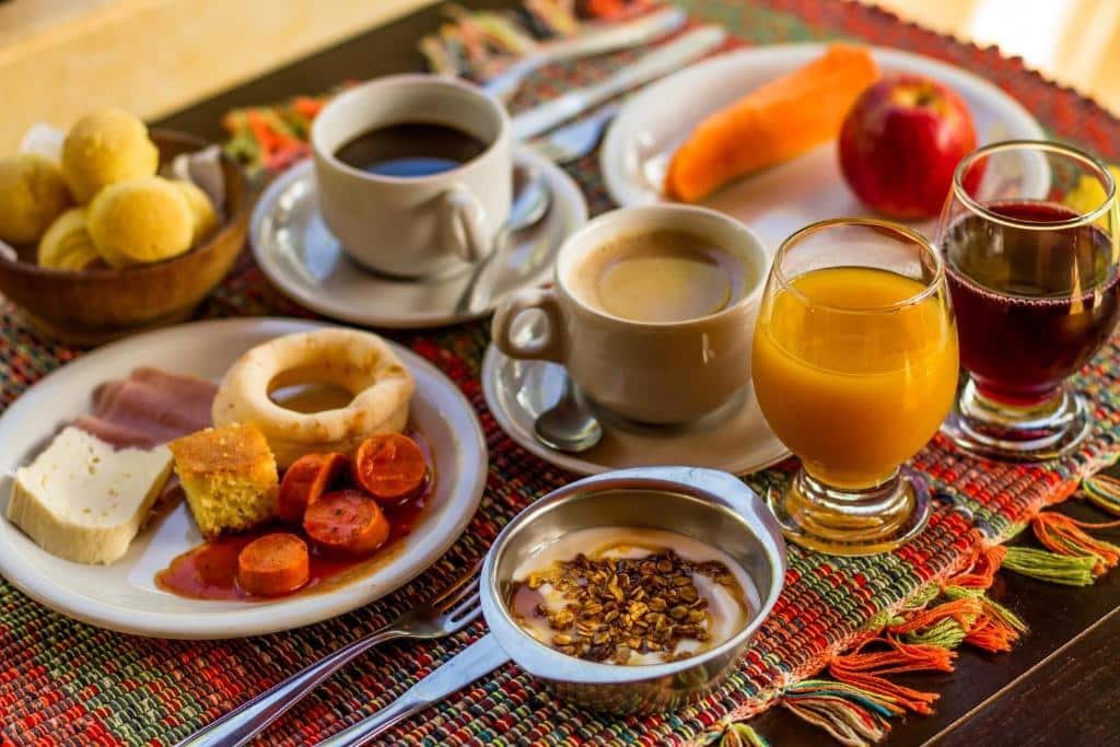 mesa de café da manhã com vários pratos, xícaras e copos com sucos. Há uma xícara com café e outra com café e leite, um prato com queijo, salsicha, bolo e polvilho, outro prato com uma maçã e pedaço de mamão, um copo de suco de laranja e outro de uva, uma cumbuca com pães de queijo e outra com iogurte e granola.