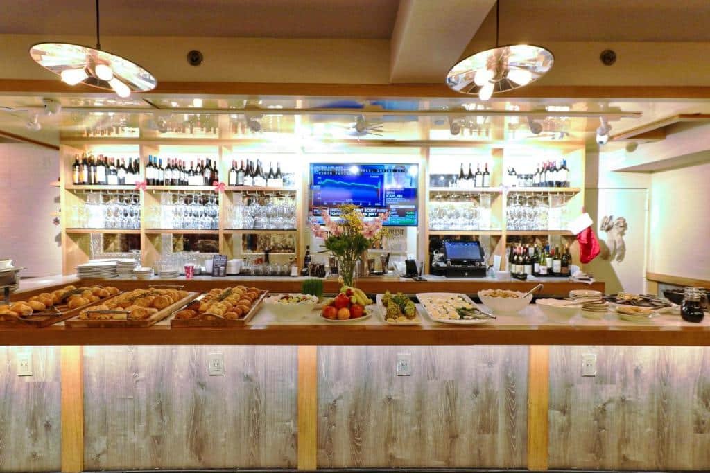 balcão de café da manhã do The Pearl Hotel com uma longa bancada de alimentos: pães, frutas, frios e geleias. Atrás da bancada é possível ver várias prateleiras de taças e garrafas de vinhos.