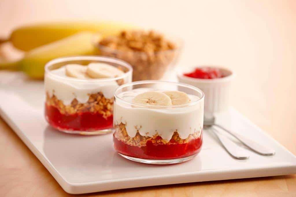 prato de porcelana quadrado e baixo com bananas, uma tigela de vidro cheia de granola, um potinho redondo de geleia de morango e duas tigelas de vidro, redondas, com um doce de banana dentro.