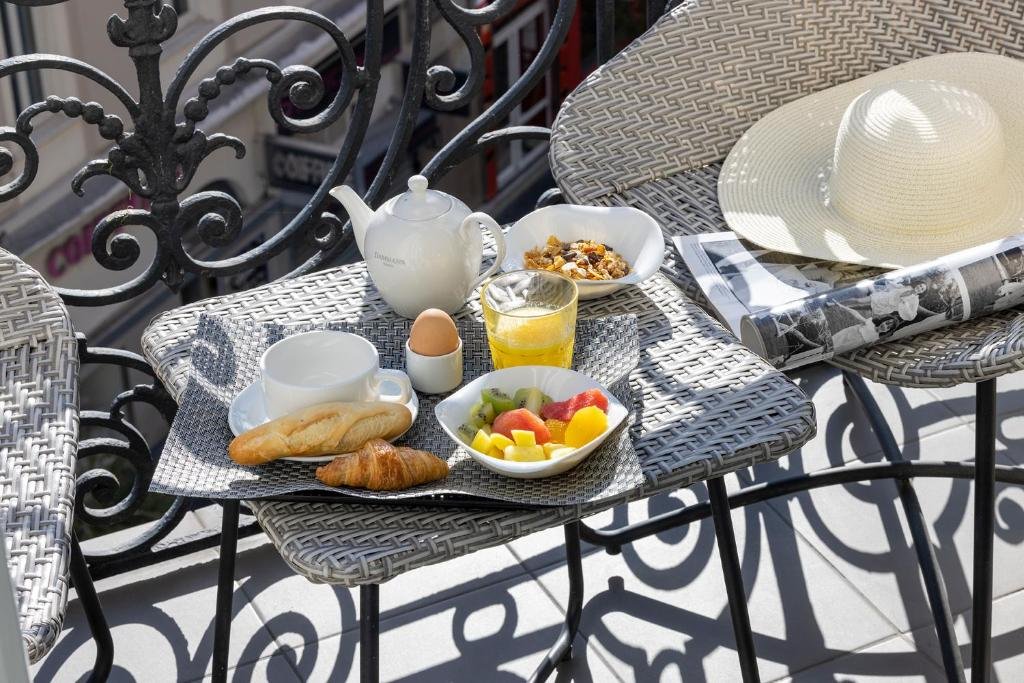 Mesa de café da manhã na varanda do Hotel 66 Nice. Em cima da pequena mesa há frutas, granola, suco, pão e café. Ao lado da mesinha há duas cadeiras, uma com uma revista e um chapéu em cima.