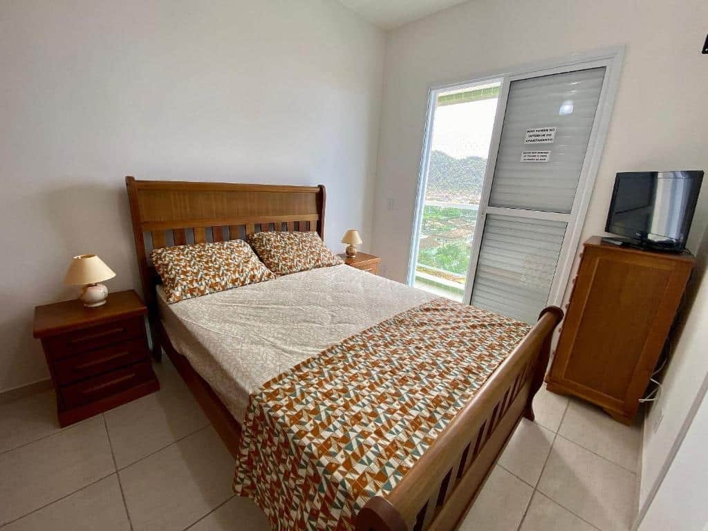 Quarto com cama de casal centralizada, pequeno móvel com luminário em cada lado da cama, porta de vidro para a varanda no lado direito e cômoda com televisão também no lado direito. Imagem para ilustrar o post pousadas em Mongaguá.