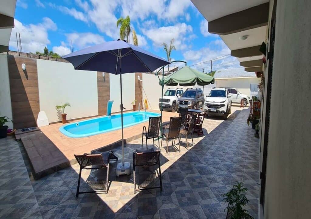 Área da piscina da Cananeia Suítes. A piscina está no canto direito, embaixo do sol. Ao seu lado esquerdo estão espreguiçadeiras e dois guarda-sóis. Ao fundo estão quatro carros estacionados.