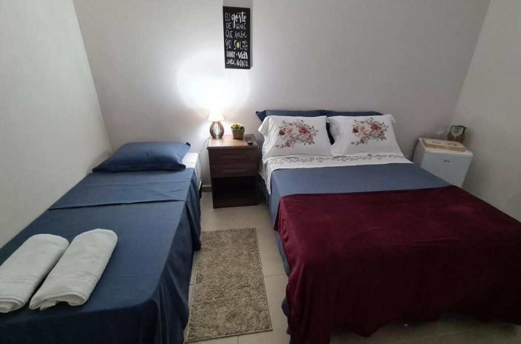 Quarto da Cananeia Suítes. Existem duas camas lado a lado, uma de casal e outra de solteiro. No meio delas há uma pequena mesinha com abajur