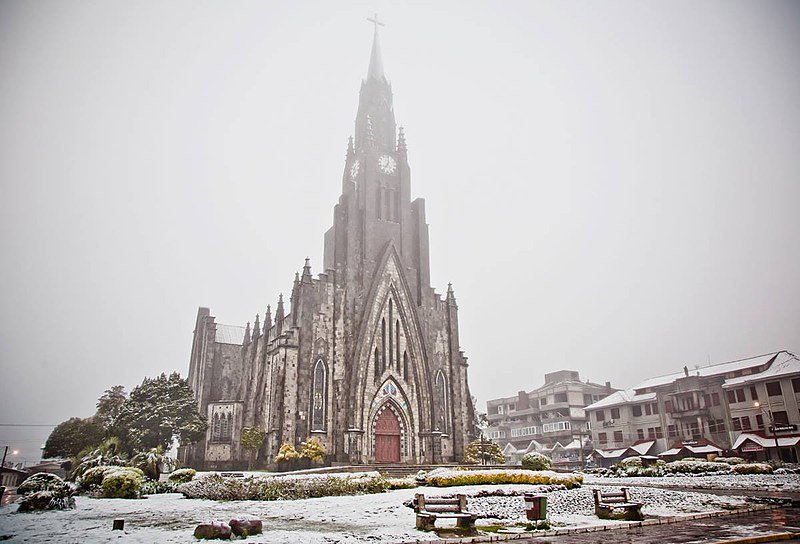 Imagem da Catedral Nossa Senhora de Lourdes, em Canela, em um dia de neve. Sua torre é alta e possui um relógio no topo. Ao seu redor há casas e arborização, também cobertos pela neve.