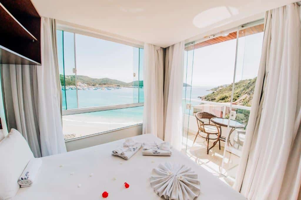 Quarto do Canto da Canoa. No canto esquerdo da imagem está a cama de casal do quarto. Já no lado direito há uma janela e uma porta para a varanda com vista total do mar. Na varanda há uma mesa redonda e duas cadeiras. Imagem utilizada para ilustrar o post airbnb em Arraial do Cabo.