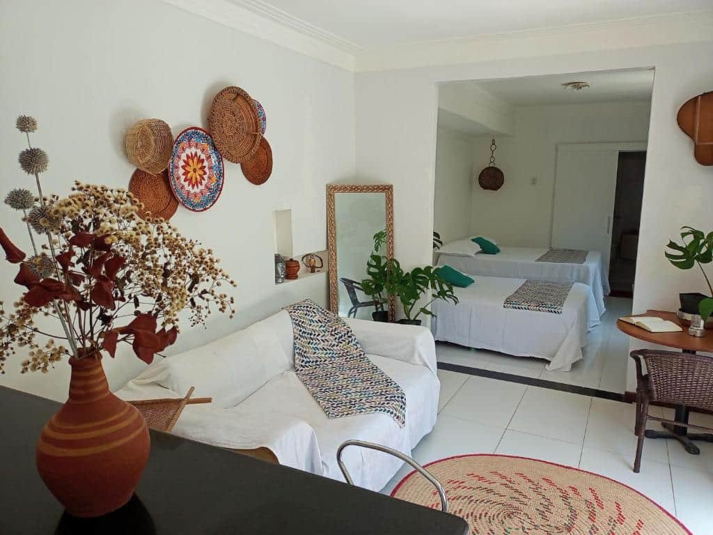 Quarto do Canto Pedra da Lua. Uma bancada na frente com um vaso de flor, um sofá no lado esquerdo com um espelho, no lado direito uma mesa com uma cadeira. No fundo, uma cama de solteiro e uma cama de casal. Foto para ilustrar post sobre pousadas em Arembepe.