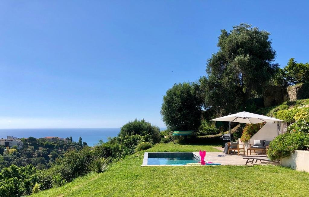Foto do quintal da casa de temporada La Coulina. No lado esquerdo há uma bela paisagem com casas em uma área verde e o mar no fundo. Já no lado direito há uma piscina na área verde da casa com pisa, cadeira e um guarda-sol. Imagem para ilustrar o post hotéis em Nice.