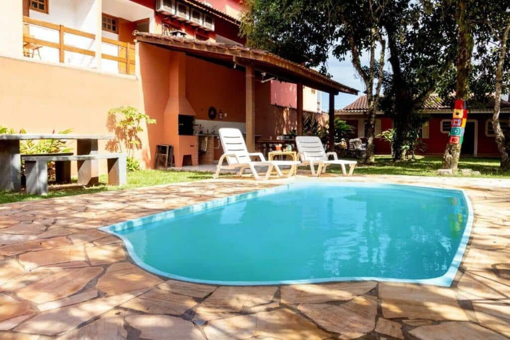 Piscina da Casa ampla c/ piscina perto da praia em Boiçucanga. Ela está no meio de uma área externa, perto de duas espreguiçadeiras. Ao fundo está a casa.