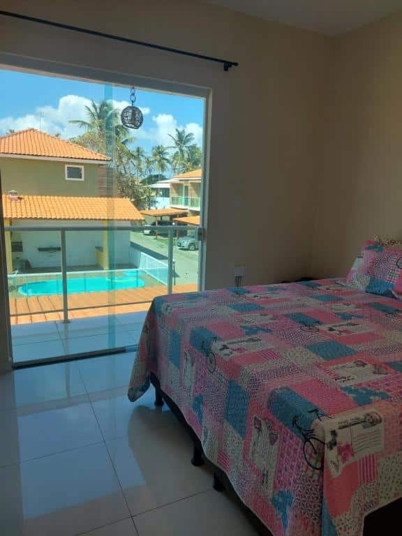 Quarto da Casa Arembepe em frente as piscinas naturais. Uma cama de casal no lado direito, atrás o vidro com a sacada e a vista para a piscina.