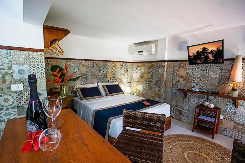 Quarto da Casa Celestino Residence. Do lado esquerdo uma bancada com taças. No fundo uma cama de casal. Do lado direito um ar-condicionado, uma prateleira e uma televisão com abajur. Foto para ilustrar post sobre airbnb em Morro de São Paulo.