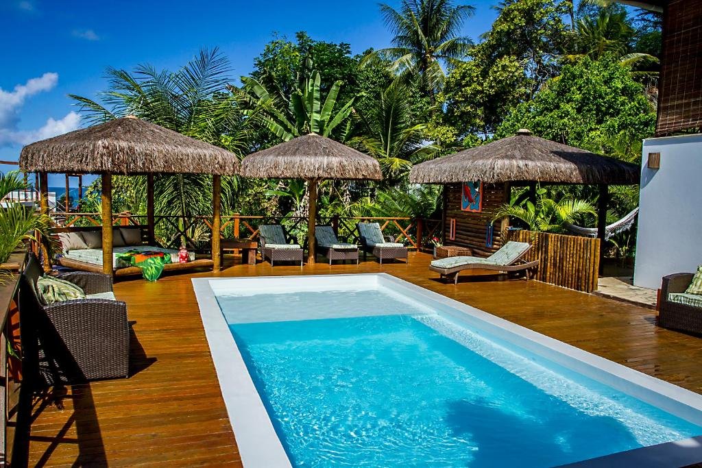 Área externa da Casa Celestino Residence. Uma piscina no meio, ao redor cadeiras de tomar sol, guarda-sol e no fundo palmeiras e a vista do mar.
