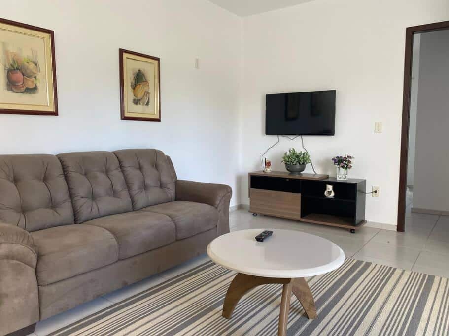 Sala de jantar da Casa Confortável em Joinville com um sofá acinzentado no lado esquerdo, uma mesa de centro redonda em frente e uma televisão presa à parede, ao fundo da sala.