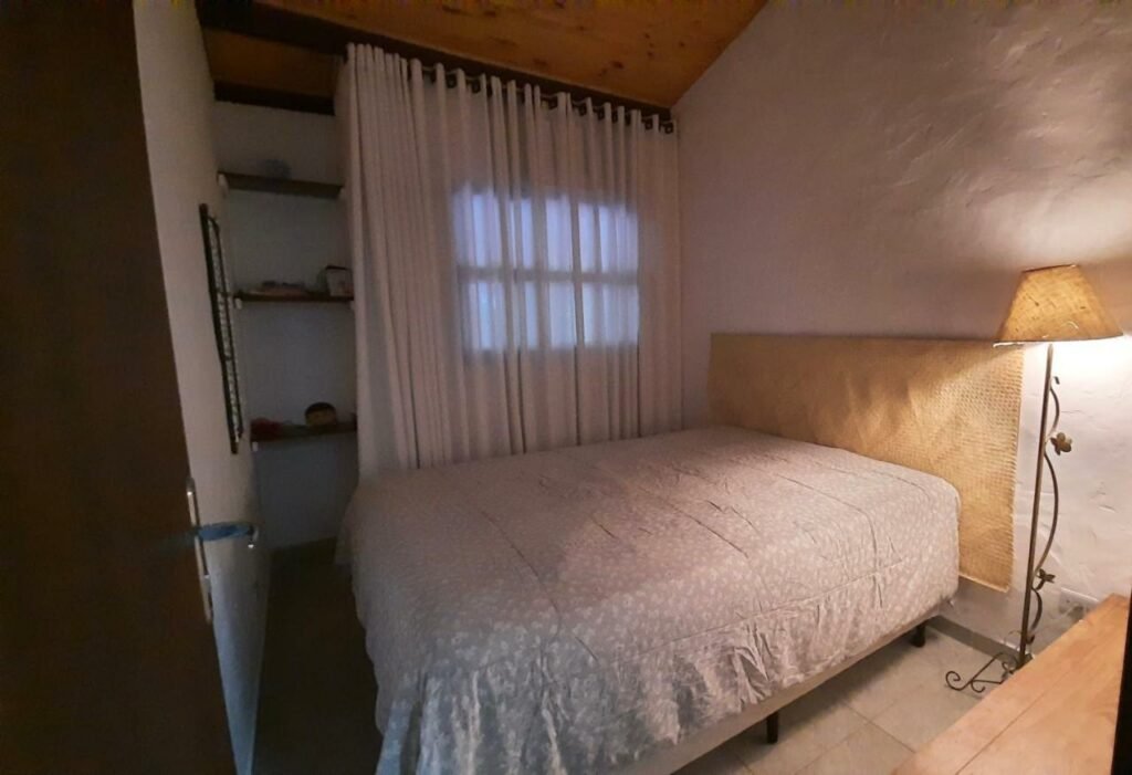 Quarto da Casa de Praia com piscina. Uma cama de casal está no meio do quaero, ao lado de uma luminária e de uma janela coberta por uma cortina. No canto direito da imagem é possível ver o pedraço de uma mesa.