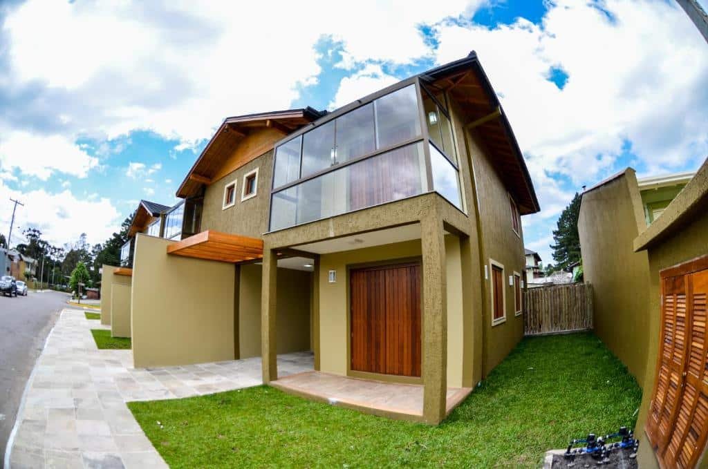 Fachada da Casas dos Alpes , uma casa com dois andares, uma sacada envidraçada, tudo em tons de verde e com detalhes em madeira, com um pequeno gramado ao redor e de frente para a rua