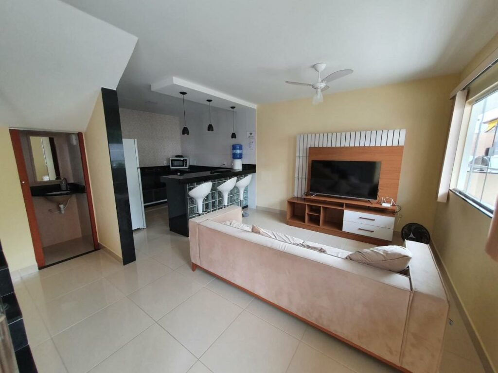 Casa Duplex Iriri - Banheira e Piscina. Um sofá na frente e um painel com uma televisão atrás. No canto direito a janela do apartamento. No lado esquerdo a porta do banheiro e do lado a cozinha, com um balcão, três cadeiras, uma geladeira e um microondas.