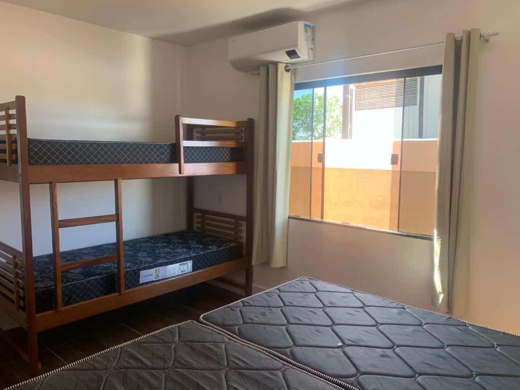 Quarto do airbnb Casa em condomínio fechado. No lado esquerdo da imagem há uma beliche, no lado direito há dois colchões de solteiros um do lado do outro formando uma cama de casal e ao fundo há uma janela de vidro com cortina. Imagem utilizada para ilustrar o post airbnb em Arraial do Cabo.