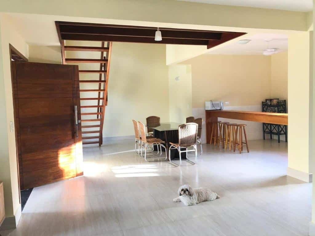 Casa em Paúba. O ambiente é aberto e espaçoso. A porta de entrada está à esquerda, um cachorrinho deita no meio da sala. No fundo há uma escada, uma mesa de vidro com cadeiras e outra bancada de madeira com alguns bancos.