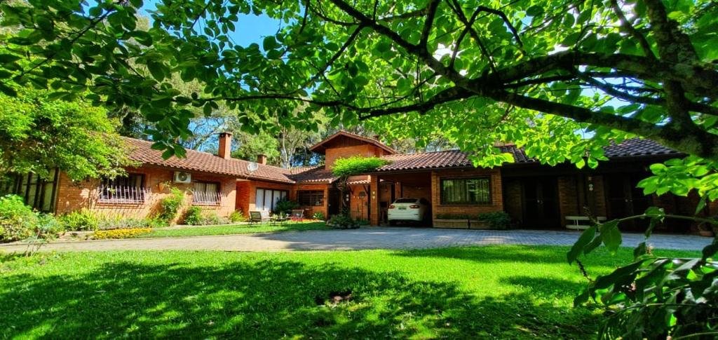 Entrada da CASA GOLDONI GRAMADO 3 Quartos, um local muito amplo e cercado por um extenso gramado e árvores, a construção é marrom e de tijolinhos