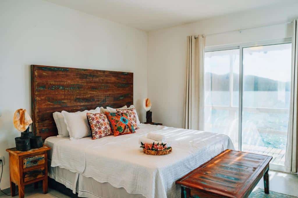 Quarto da Casa Grega. A cama de casal está posicionada no lado esquerdo do quarto e em cada lado da cama há mesa de cabeceira. Em frente a cama há um banco de madeira. No fundo há uma porta de vidro que dá acesso a varanda e também oferece vista para o mar. Imagem utilizada para ilustrar o post airbnb em Arraial do Cabo.