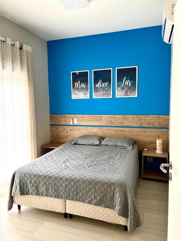 Foto do quarto no Residencial Marina Del Sol. Há uma cama de casal no centro, e atrás há uma cabeceira de madeira e três quadros. Na esquerda há uma janela com cortinas.