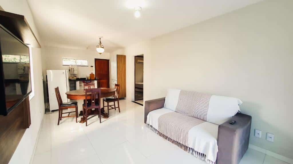 CASA NA RUA DO PIRUÍ EM AREMBEPE com ar. Um sofá no lado direito, de frente uma televisão. No fundo, uma mesa com quatro cadeiras, a cozinha, geladeira e a porta.